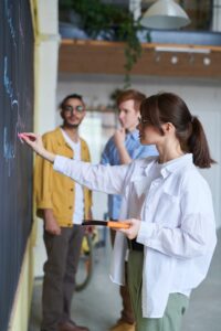 Kobieta pisze kredą po tablicy w klasie na lekcji angielskiego, a w tle dwóch mężczyzn.